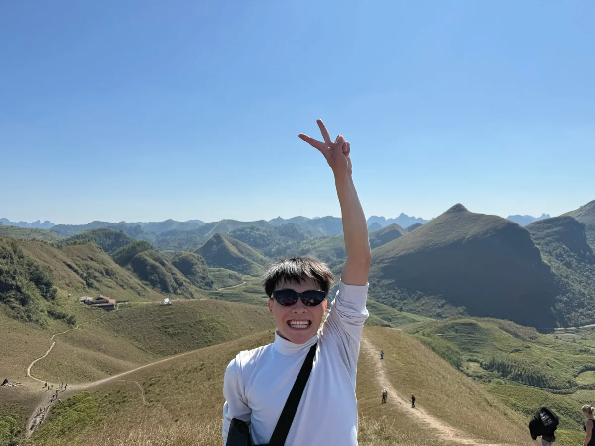 Person in sunglasses, smiling with a peace sign in front of green mountains under a clear blue sky, exuding a joyful mood.