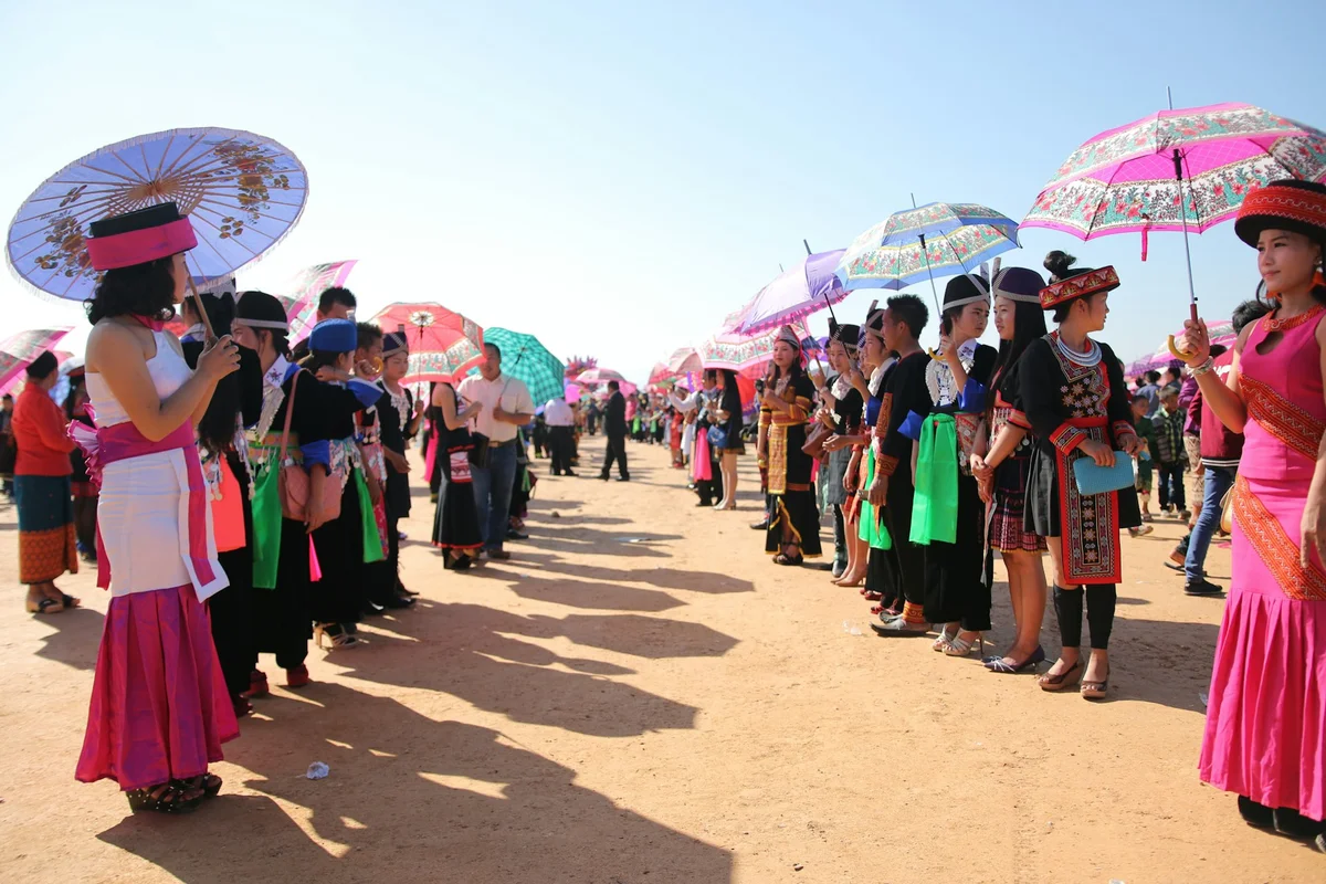 People in colorful traditional clothing and vibrant umbrellas stand in two lines on a sunny day, creating a festive atmosphere.