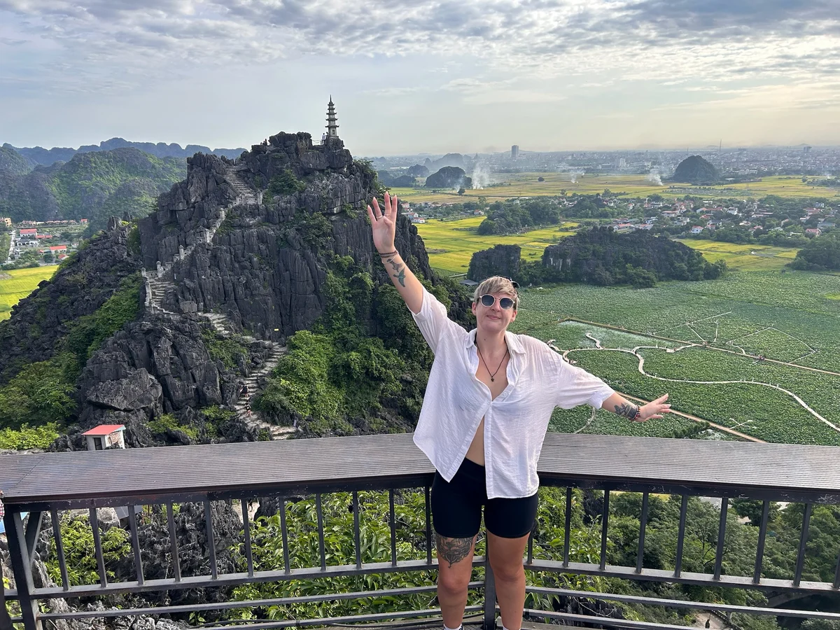 Person in sunglasses raising arms on a viewpoint with rocky hill, stairs, and a distant pagoda. Lush green landscape under cloudy sky.