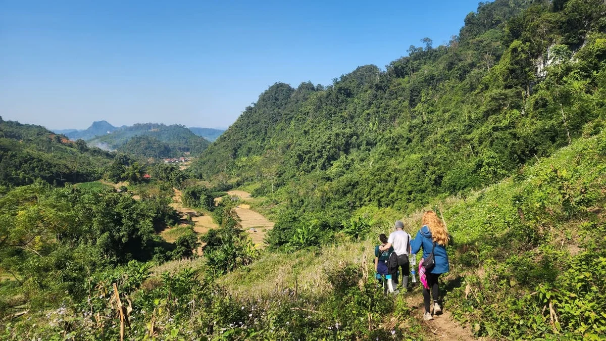 People hiking along a lush green mountain trail under a clear blue sky, surrounded by rolling hills and valleys in the distance.