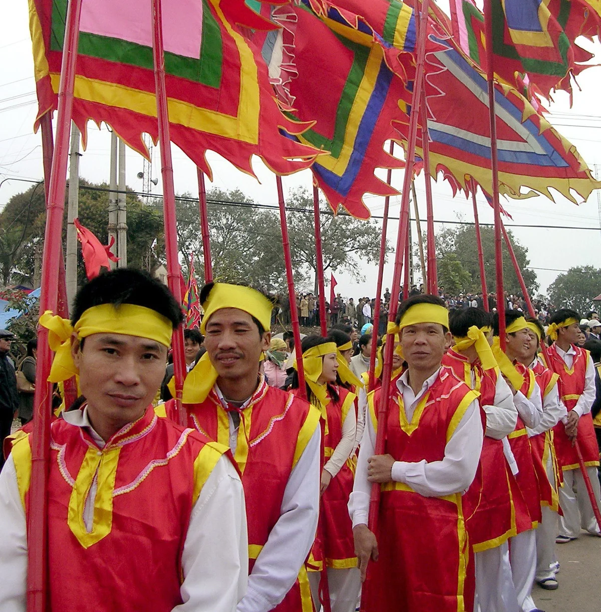 People in red and yellow traditional attire hold colorful flags during a festive outdoor event. Crowd and trees in the background.