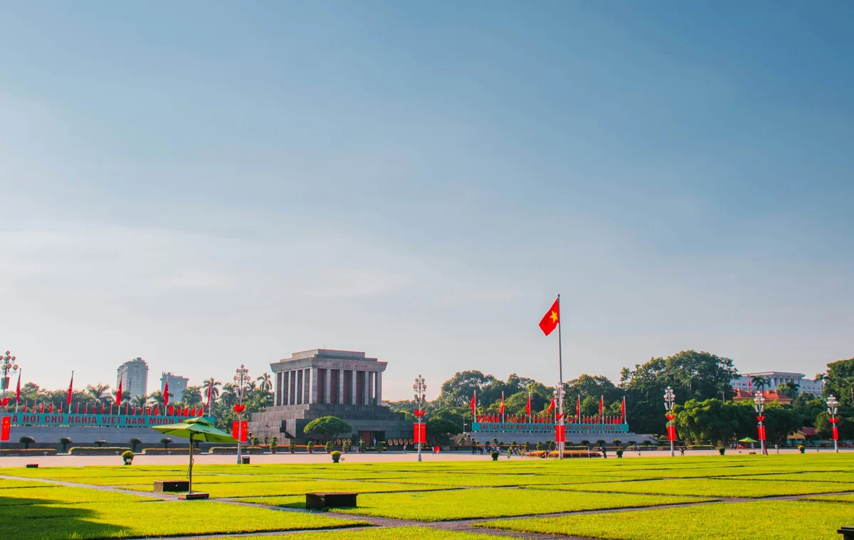 Vietnamese flag flies in front of Ho Chi Minh Mausoleum. Green lawn in foreground, blue sky. Red banners and text in background.