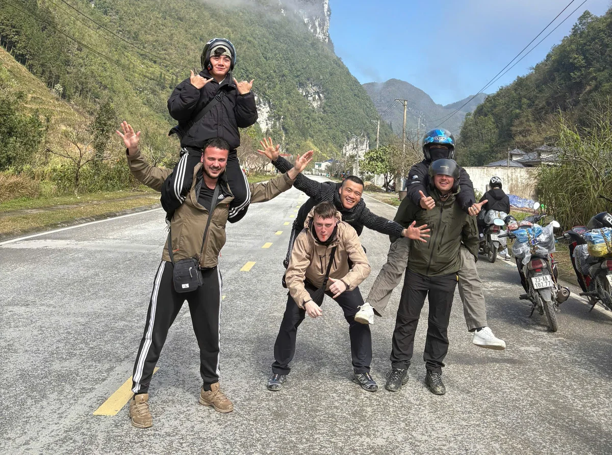 Group of six friends posing playfully on a mountain road. Two are on shoulders, wearing helmets. Mood is joyful, surrounded by greenery.