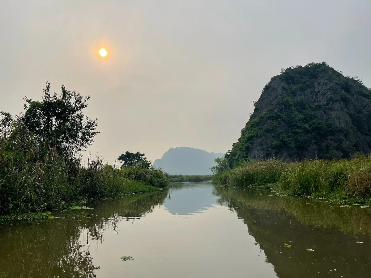 River scene with calm water reflecting a cloudy sky, surrounded by lush greenery and rocky hills. Sun is partially visible. Peaceful mood.