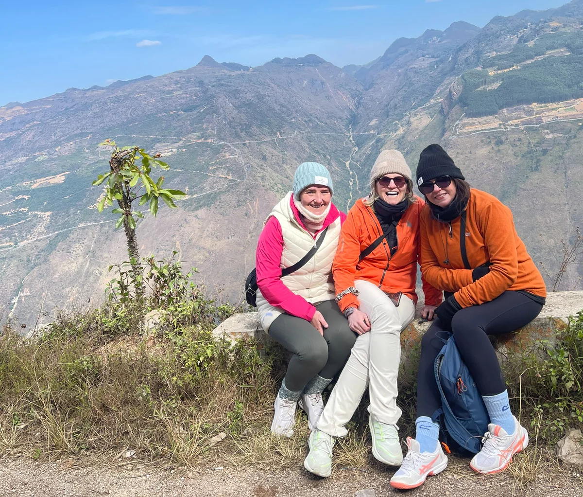 Three women in warm clothes sit smiling on a mountain ledge, with scenic hills in the background under a clear blue sky.