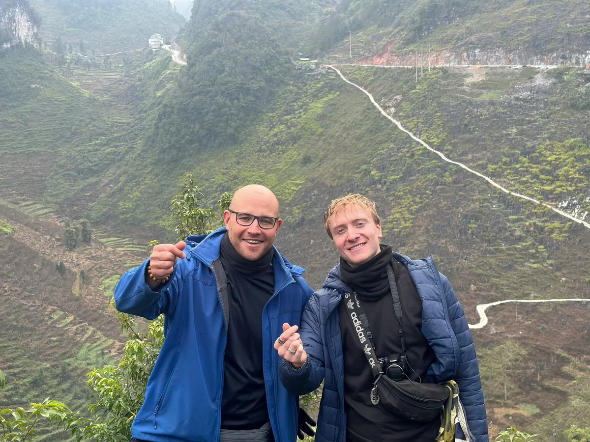 Two smiling individuals pose on a misty hillside with terraced fields and winding paths in the background, wearing blue jackets.