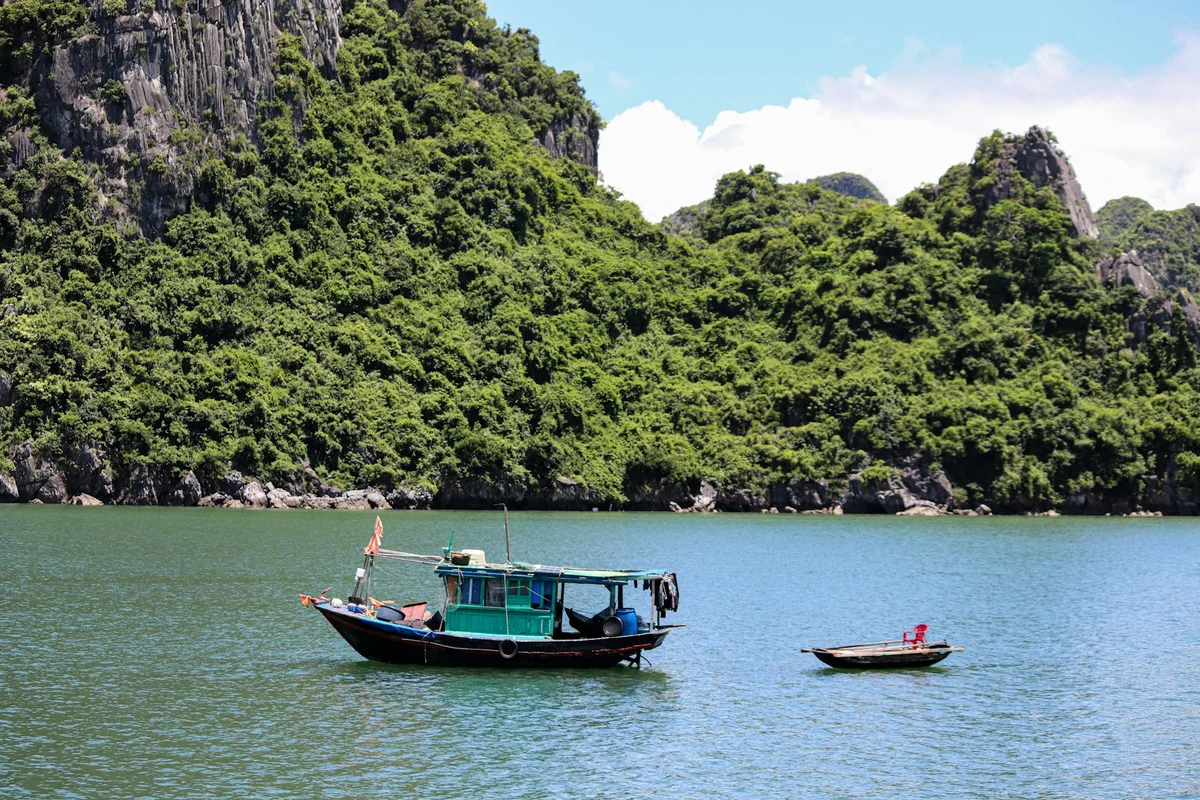 Boat in a tranquil bay with a lush green mountain backdrop. Clear blue sky and water. Small raft nearby with a red chair.