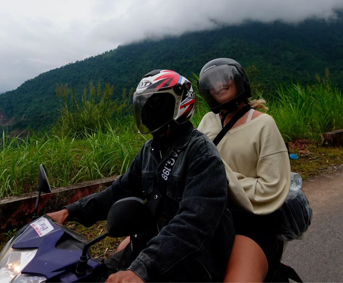 Two people wearing helmets ride a motorbike on a road with lush greenery and misty hills in the background. The mood is adventurous.