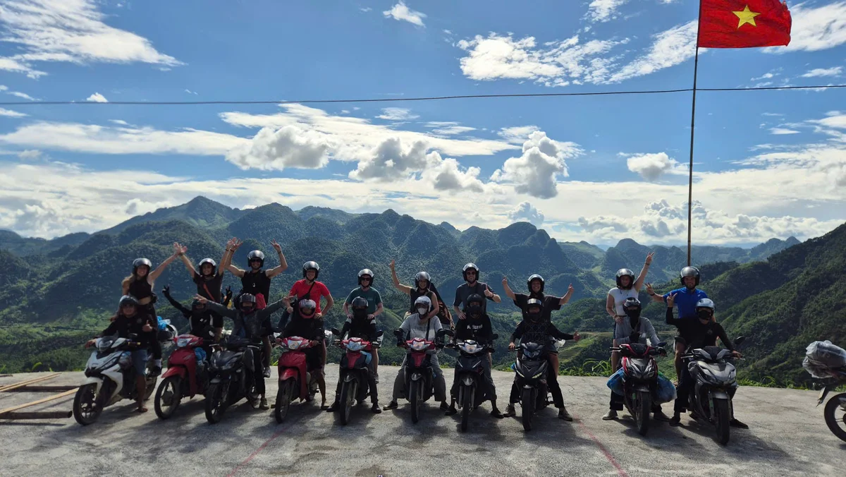 A group of people on motorcycles joyfully pose against a mountainous backdrop under a blue sky. A red flag with a yellow star waves.