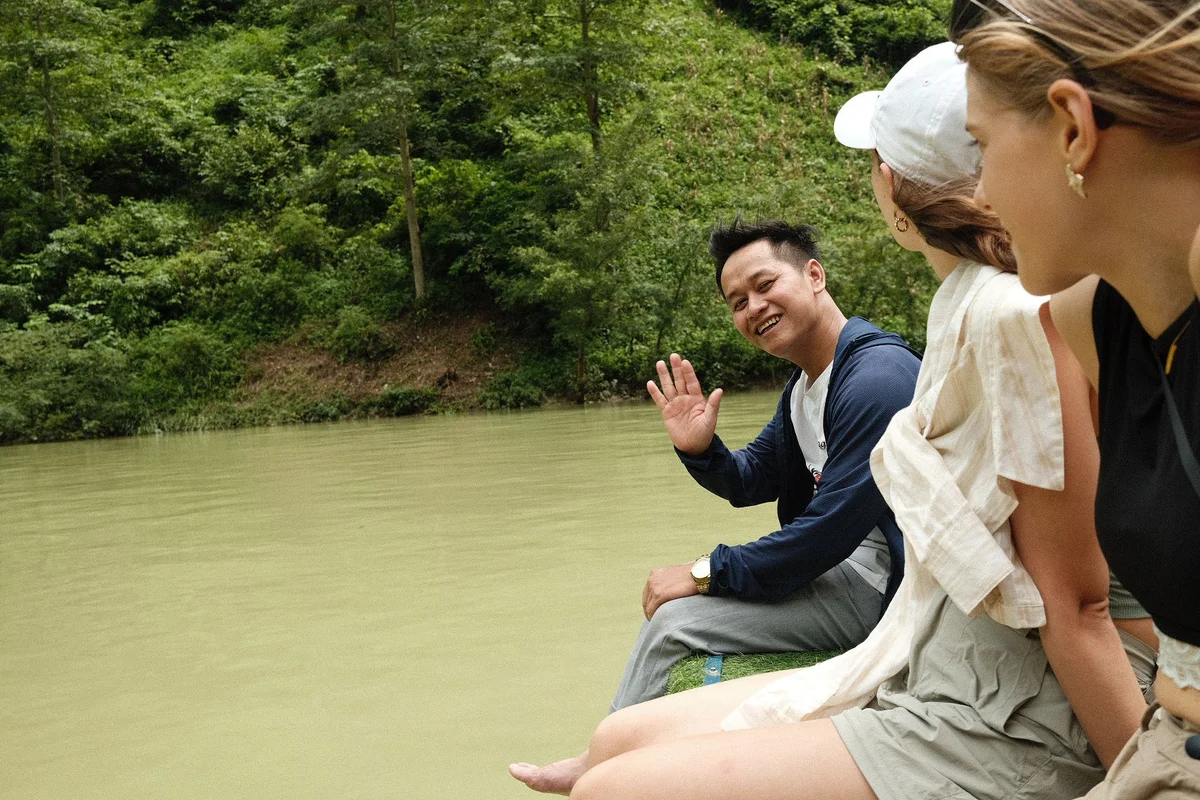 Three people sitting by a river, with one man waving and smiling. Thick green forest in the background. Casual, relaxed atmosphere.