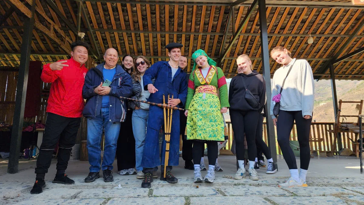 A group of smiling people pose in a wooden shelter. One wears traditional colorful attire. Sunny day with mountains visible in the background.