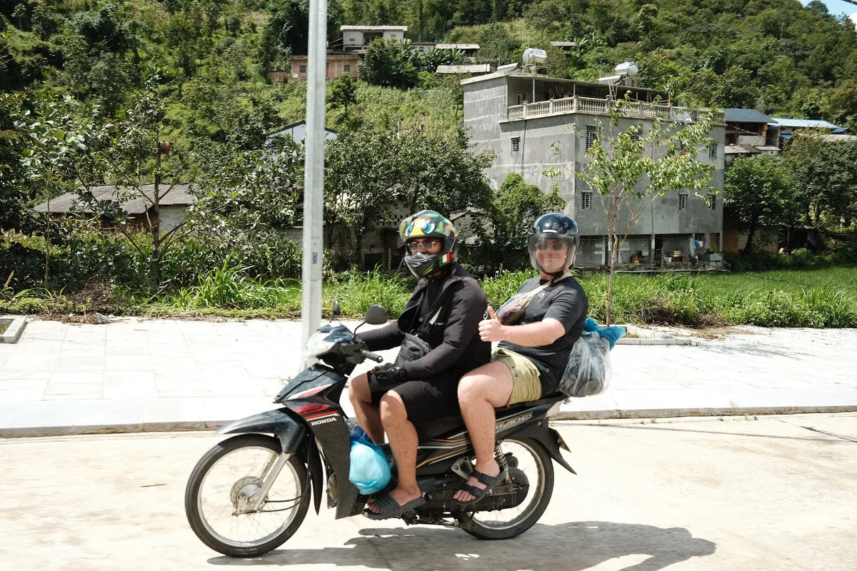 Two people on a motorcycle, both wearing helmets. One gives a thumbs up. Background: greenery and a building. Bright, sunny day.