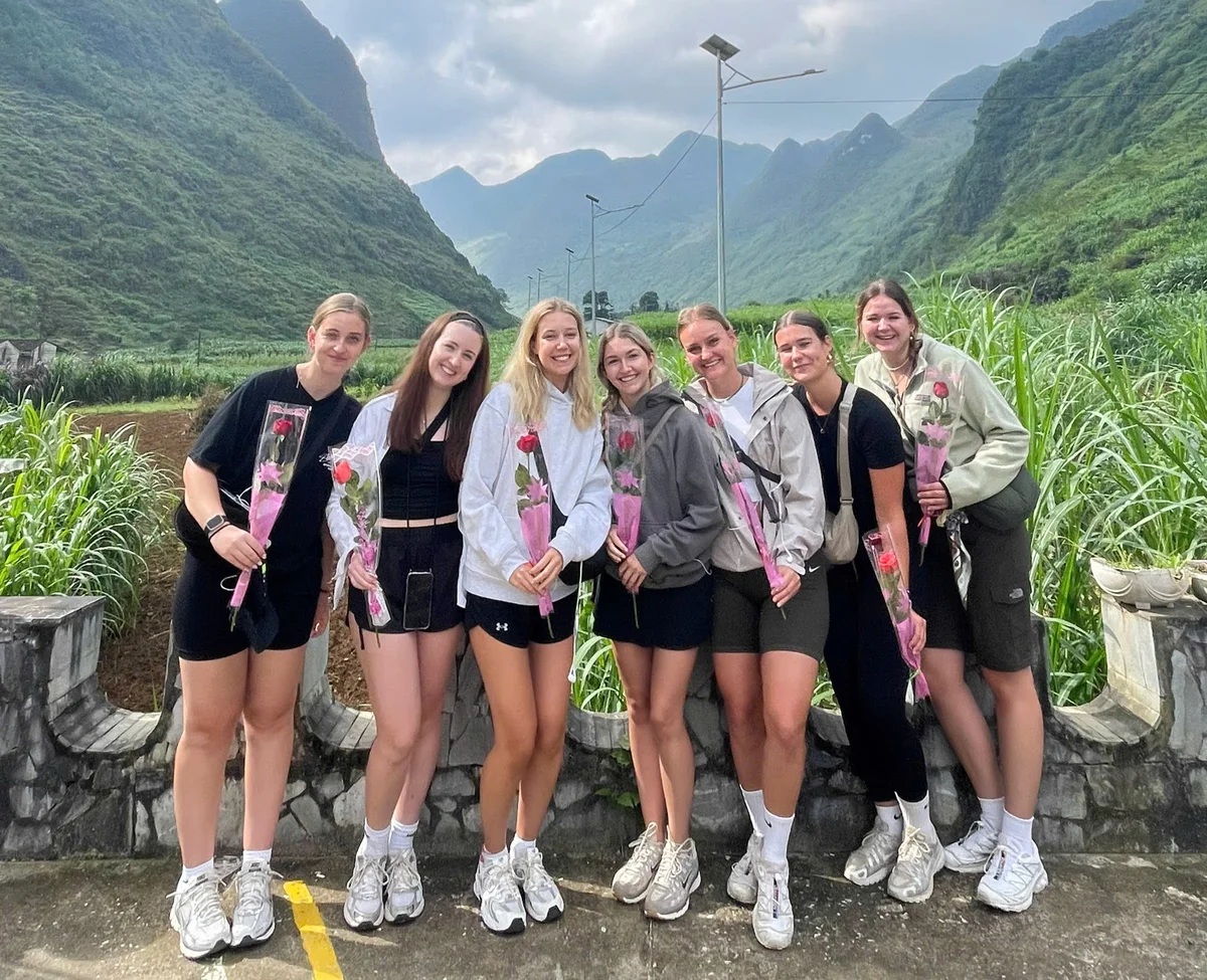 Seven people smile, holding pink-wrapped roses, standing on a stone railing with lush green hills and cloudy sky in the background.