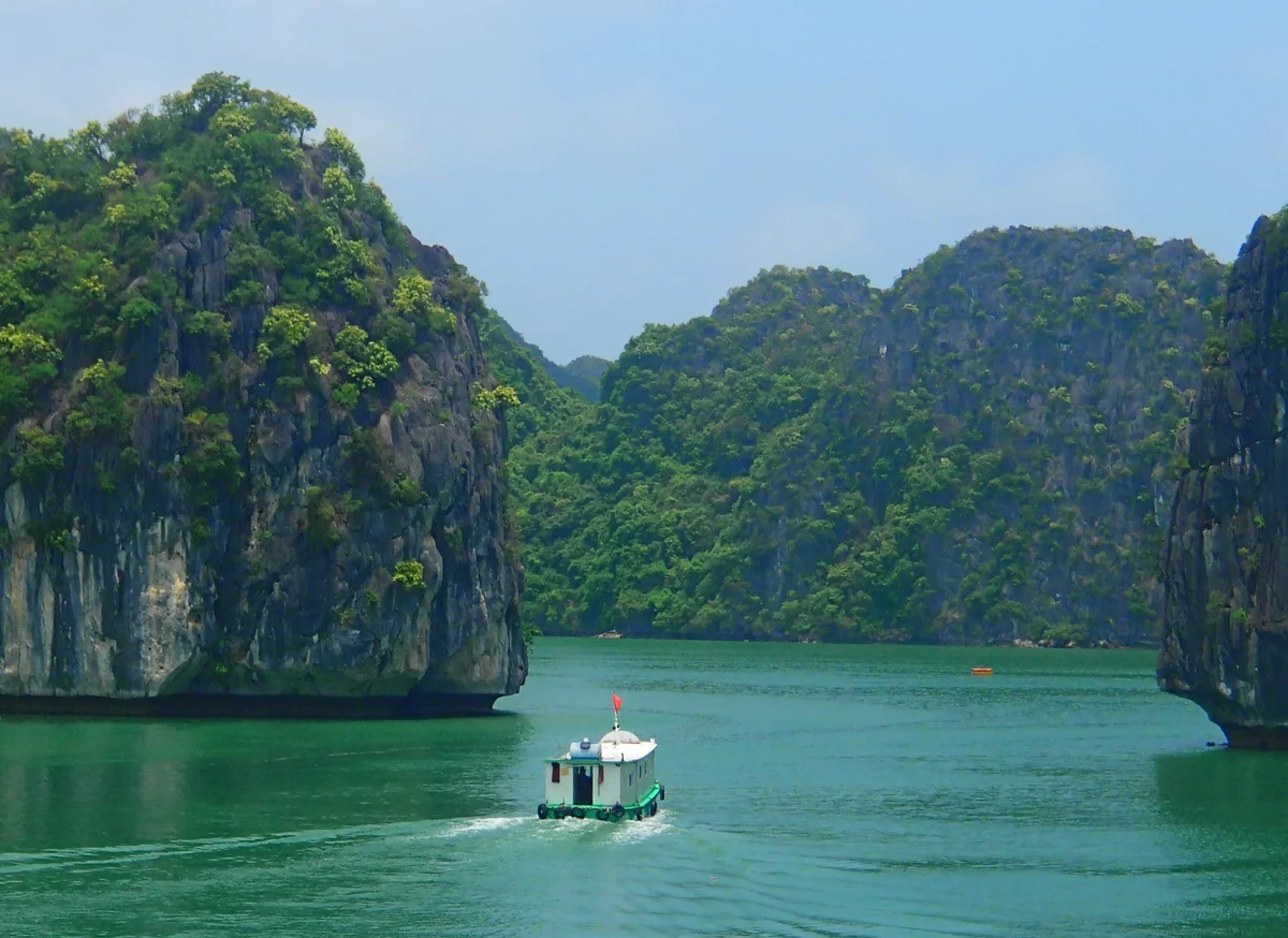 Boat with red flag sails on green water between lush, rocky cliffs under blue sky, creating a serene and peaceful scene.