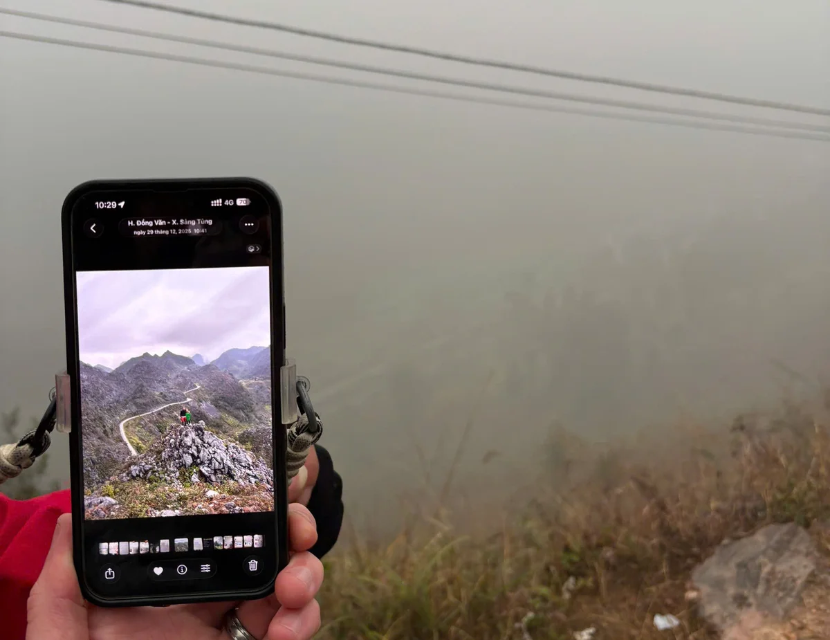 Smartphone showing a mountain trail photo held against a foggy landscape. Screen displays mountains, hikers, and app interface. Moody ambiance.