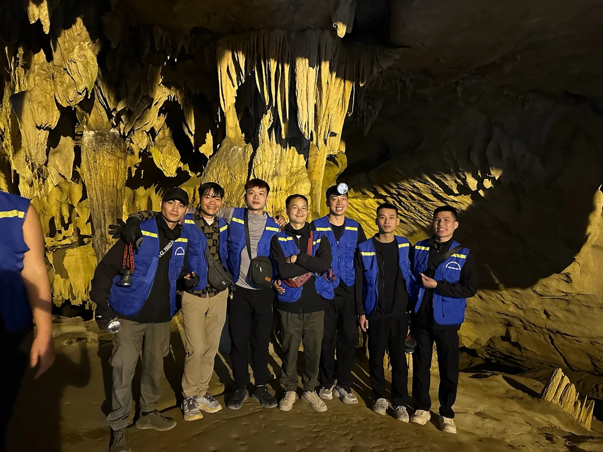 Group of seven people in blue vests smiling inside a cave with stalactites. Dim lighting gives a warm, adventurous ambiance.