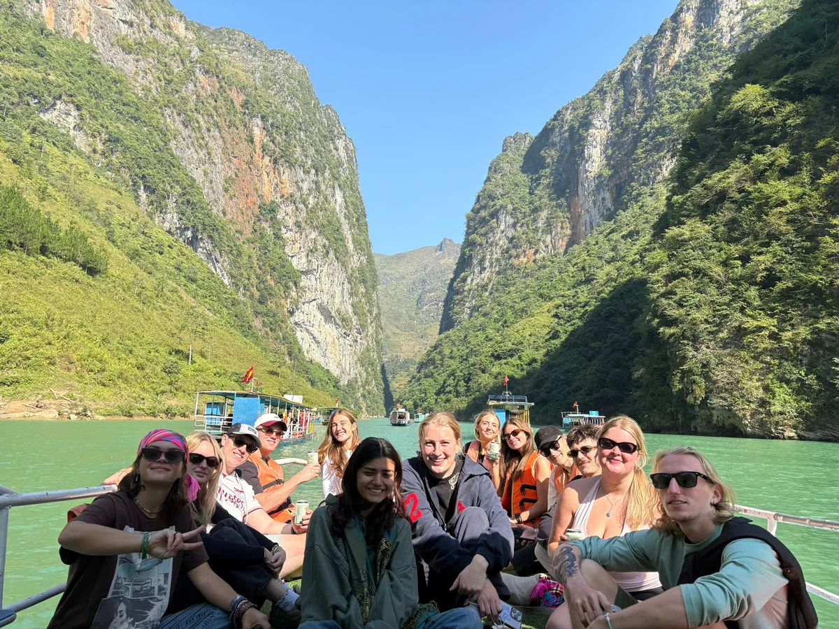 Group smiling on a boat in a scenic canyon with green water and towering cliffs. Bright, sunny day with lush vegetation around.