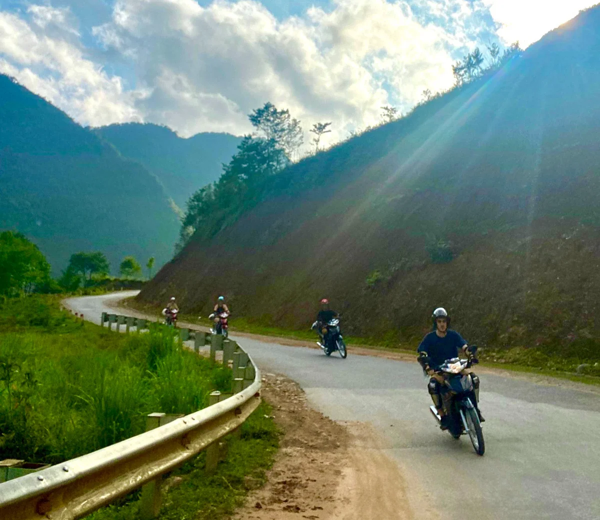 Motorcyclists ride on a winding mountain road under a blue sky with scattered clouds. Sunlight casts rays over the green landscape.