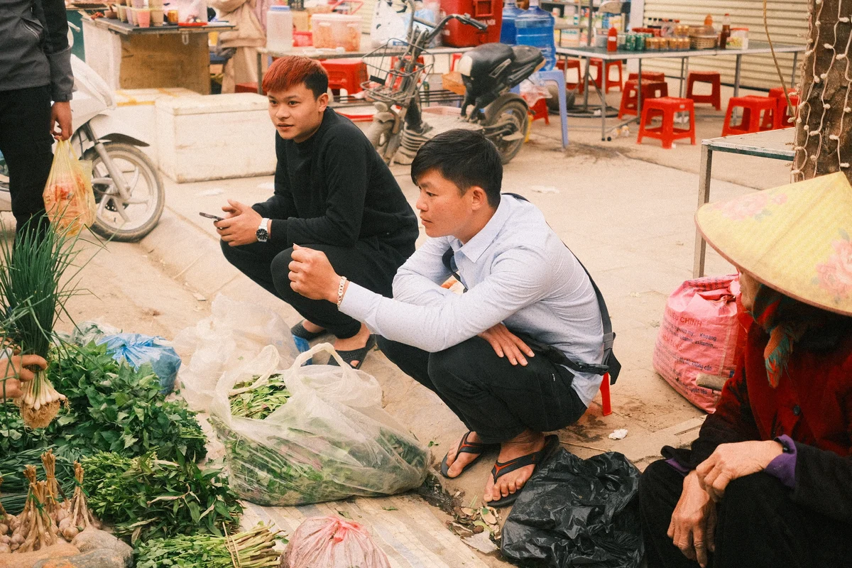 Two men squat in a market, examining vegetables in plastic bags. A woman in a conical hat sits nearby. Red chairs and bikes in background.