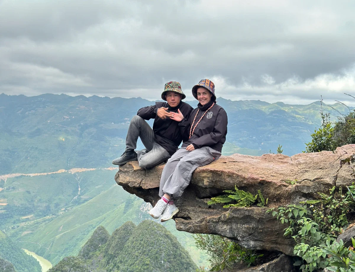 Two people sit on a cliff edge with scenic mountains in the background, wearing hats and dark jackets, appearing content and adventurous.