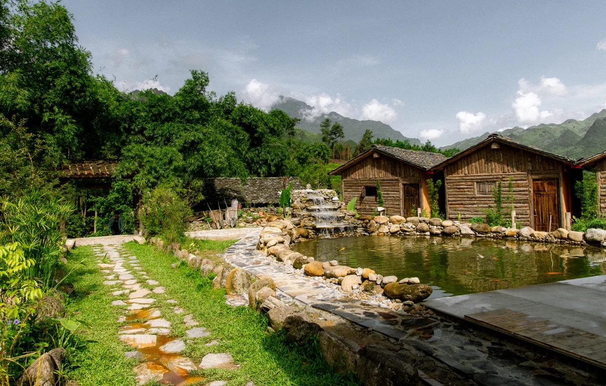 Rustic cabins by a pond with a stone waterfall, lush greenery, mountains in the background, and a stone path on a sunny day.