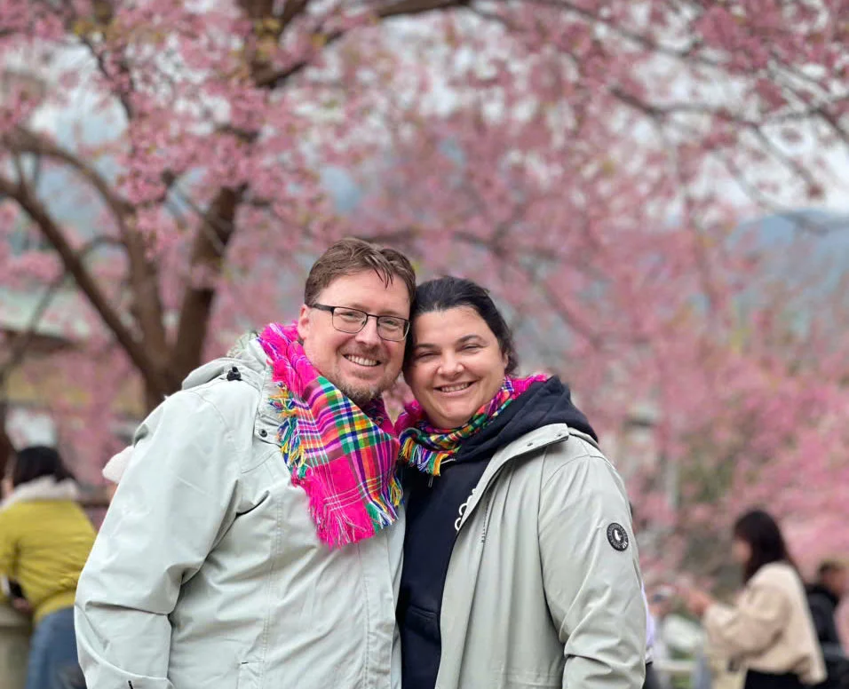 Couple smiling under pink cherry blossoms, wearing colorful scarves and jackets. People in the background add to the joyful spring atmosphere.