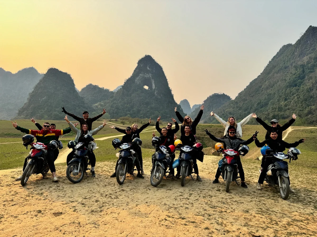 A group of people on motorbikes in a scenic landscape with mountains, holding their arms up joyfully. Dusty road in foreground, clear sky.