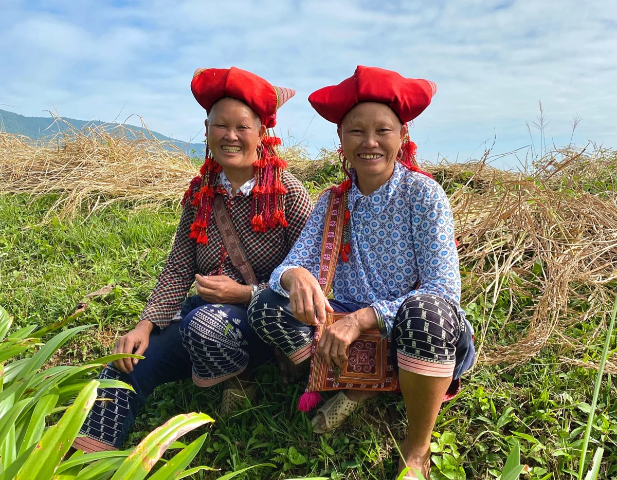 Two people in patterned clothing and red hats smile while sitting on green grass, with dry grass and distant mountains in the background.