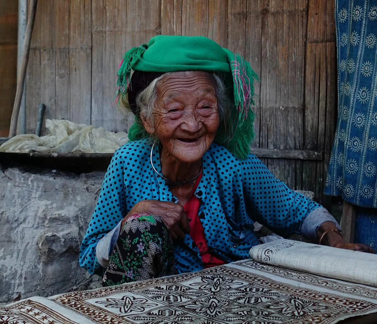 Smiling elderly woman in a green headscarf and blue polka-dot shirt, sitting beside a patterned cloth in a rustic wooden setting.