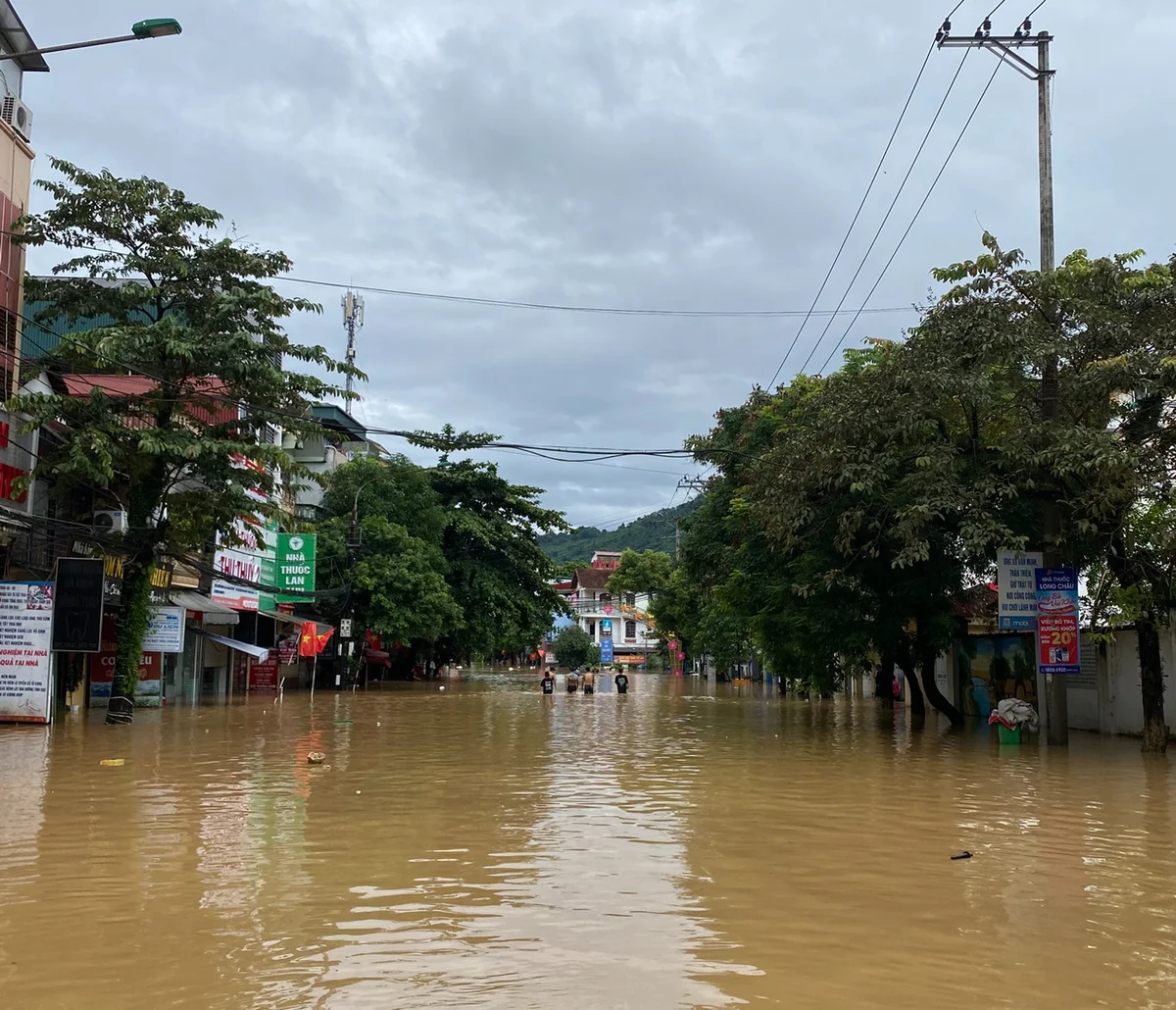 Flooded street with brown water, people wading, surrounded by trees and shops; cloudy sky, a green sign visible. Mood is tense and calm.