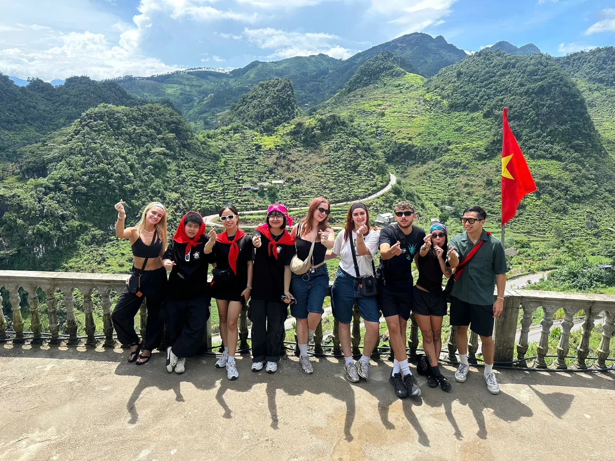 Group of people posing with hand gestures on a scenic mountain viewpoint with lush green hills, blue sky, and a Vietnam flag in the background.
