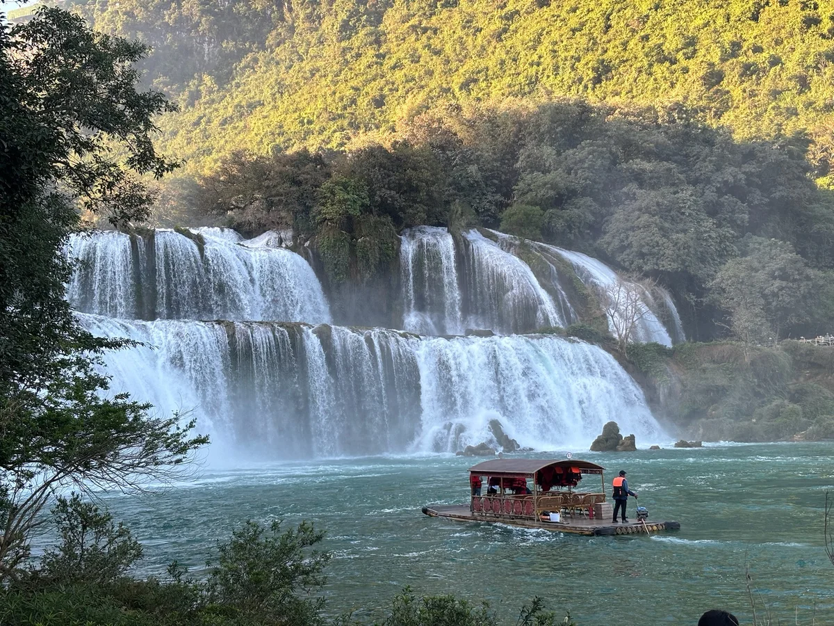 A scenic waterfall flows into a river, surrounded by lush greenery. A person in an orange vest steers a wooden raft on the water.