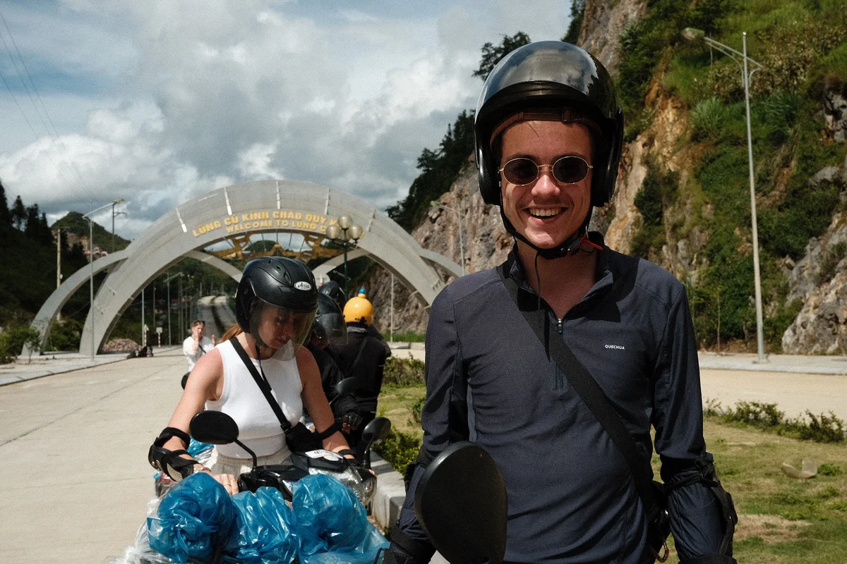 Two people in helmets on motorbikes smile under a "Welcome to Lung Cu" arch. Mountains and cloudy sky in the background, vibrant mood.