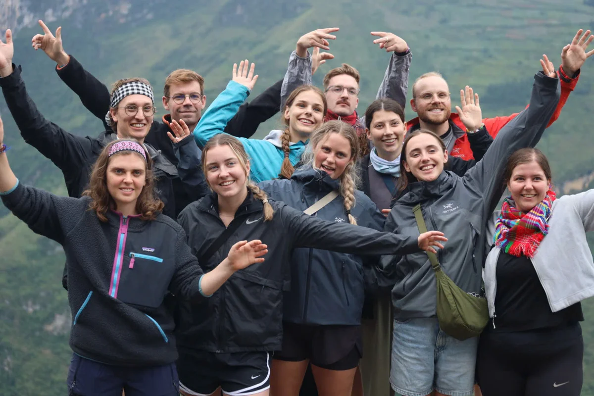 Group of smiling people posing with raised arms in a scenic outdoor setting. They're wearing colorful jackets and seem joyful and energetic.