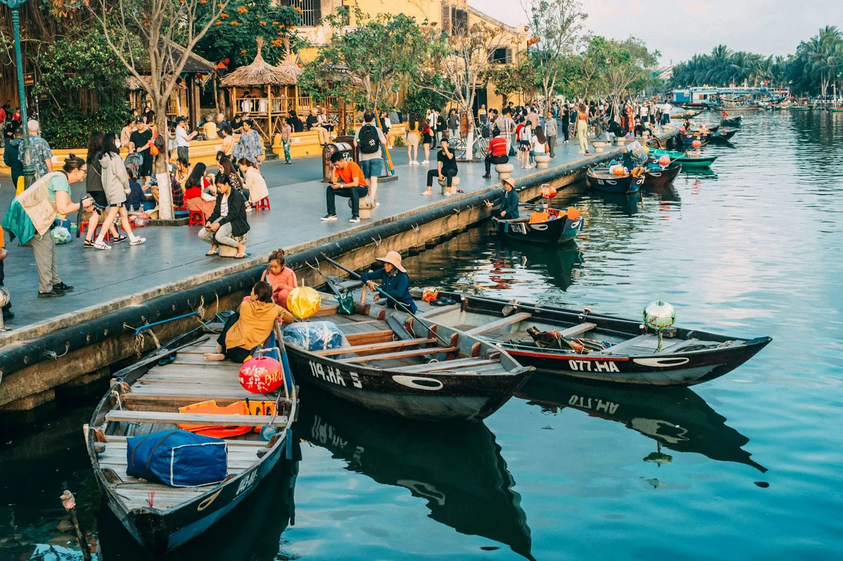 People stroll and relax along a riverside promenade in a bustling area with small boats. Lanterns add color and trees line the path.