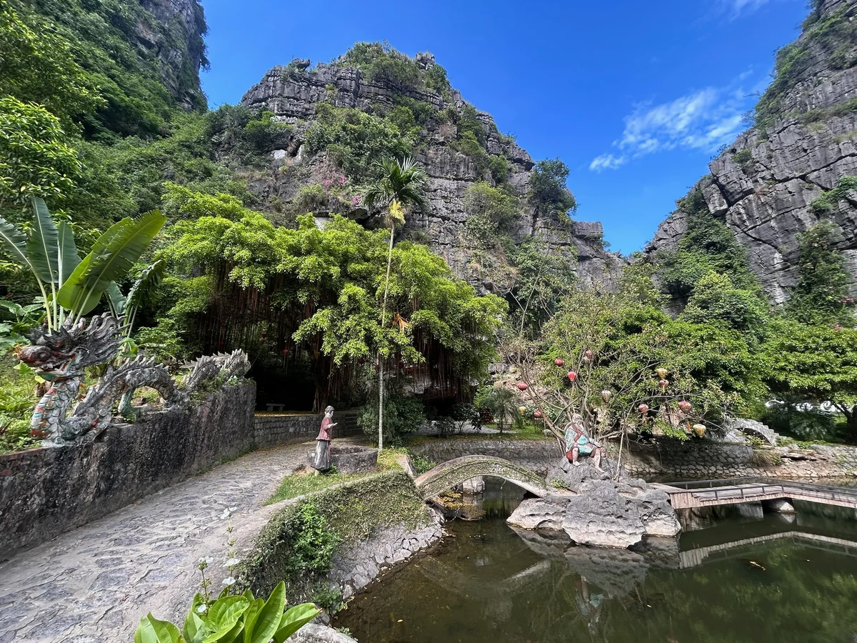 Lush mountain scene with stone dragon sculpture, a person near a rocky path, green foliage, clear blue sky, and reflective pond.