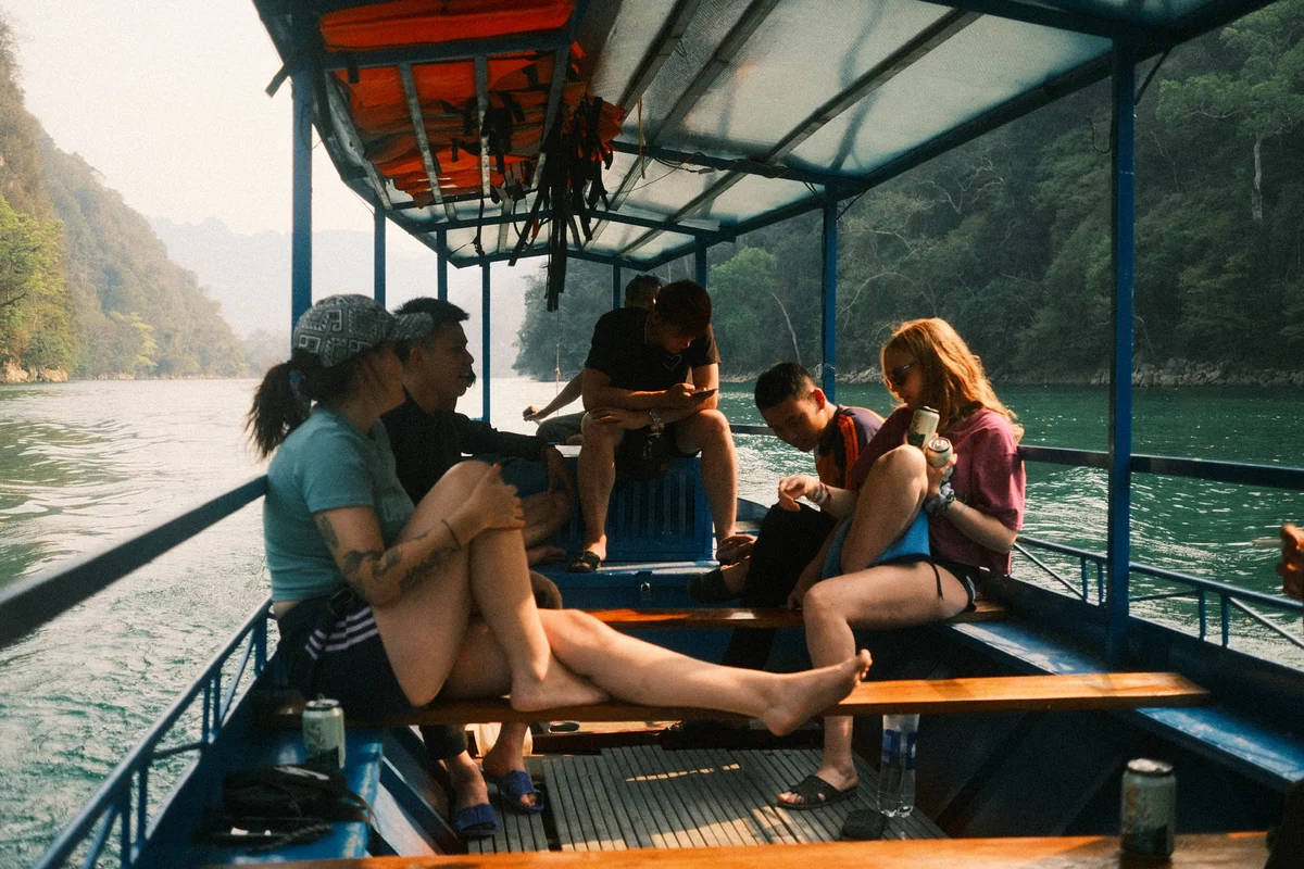 Five people relax on a boat on a green river, surrounded by forested hills. They chat, check phones, and hold drinks. A peaceful scene.