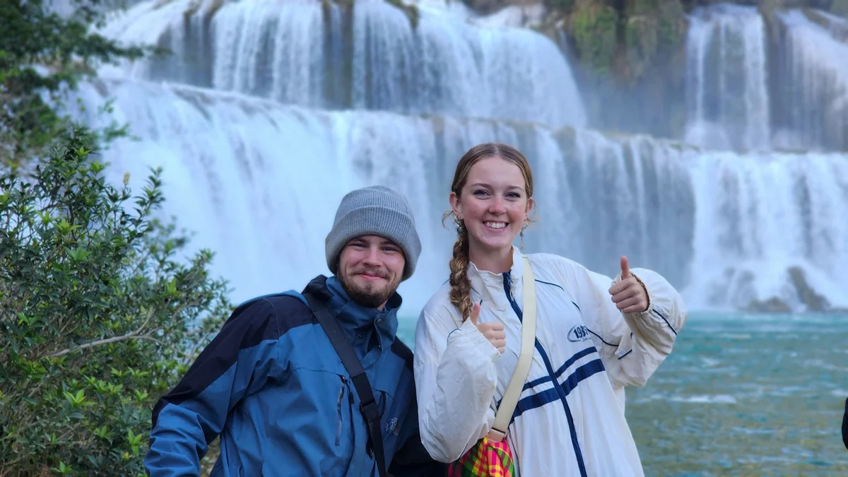 Two people smiling and giving thumbs up in front of a large waterfall. Both wear jackets; lush greenery surrounds them. Energetic and joyful mood.