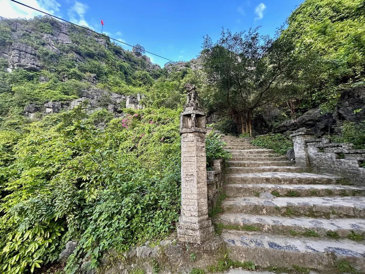 Stone steps lead up through lush greenery with a stone pillar and a sculpture. A red flag flies atop the rocky hill under a blue sky.