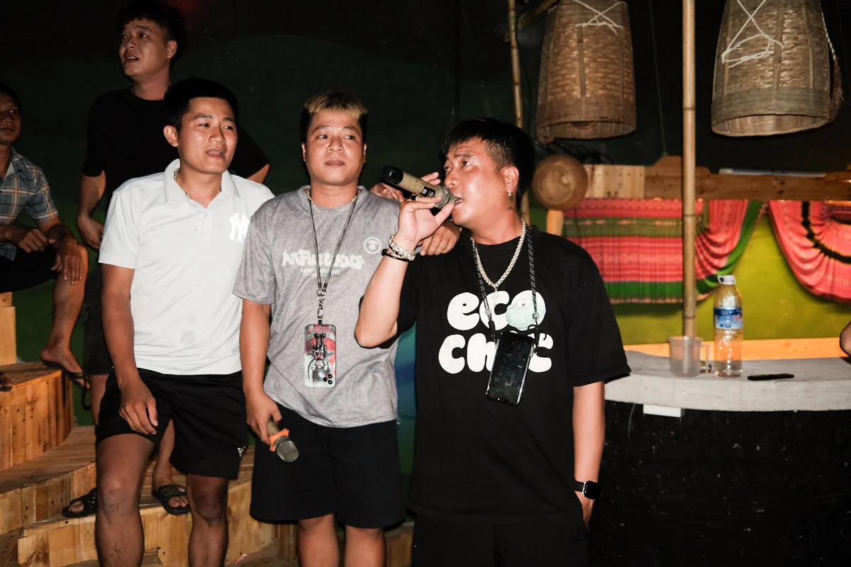 Three men are holding microphones on stage, one singing with focus. Colorful draped background, bamboo decor, and a water bottle visible.