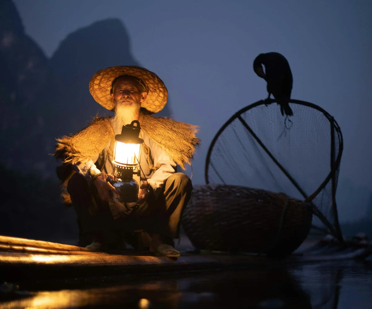 Elderly man in straw hat holds lantern on a bamboo raft at dusk, alongside a perched cormorant. Mountain silhouettes in the background.