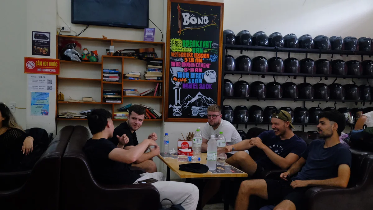 Five men sit smiling around a table in a cozy room with bookshelves and a wall of black helmets. A chalkboard lists hostel activities.