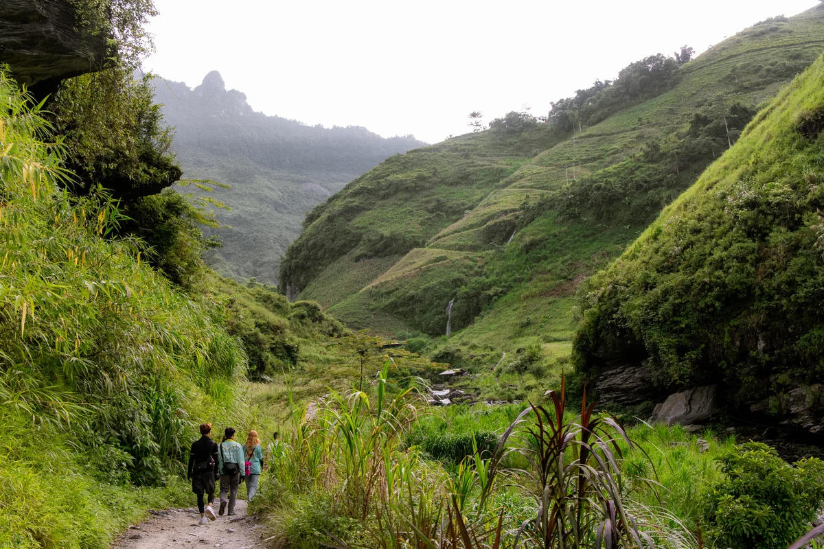 Three people walk on a lush green mountain trail with cascading hills and a small waterfall in the background. Overcast and serene setting.