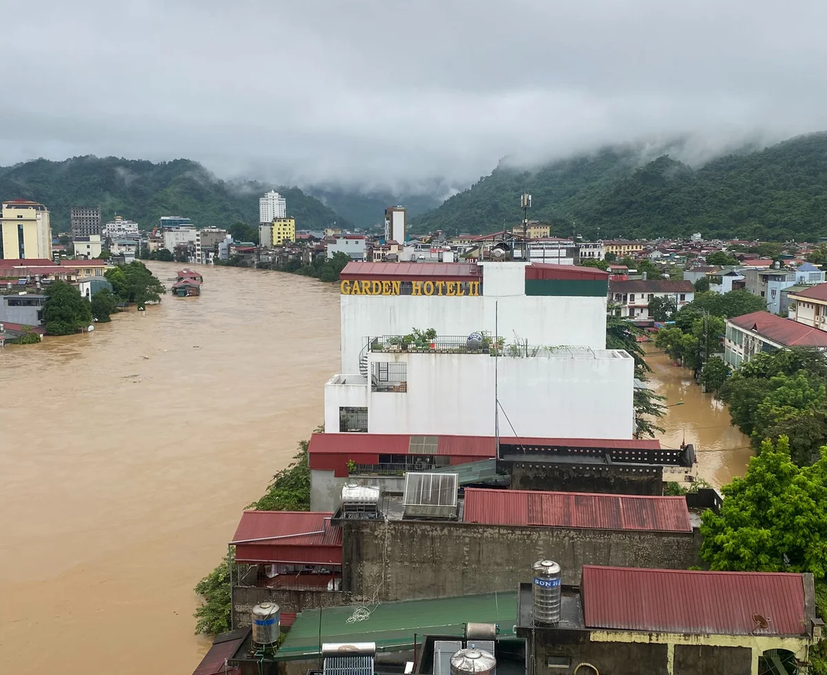 Flooded city with muddy water, buildings partially submerged. "Garden Hotel" sign visible. Overcast sky and green mountains in background.