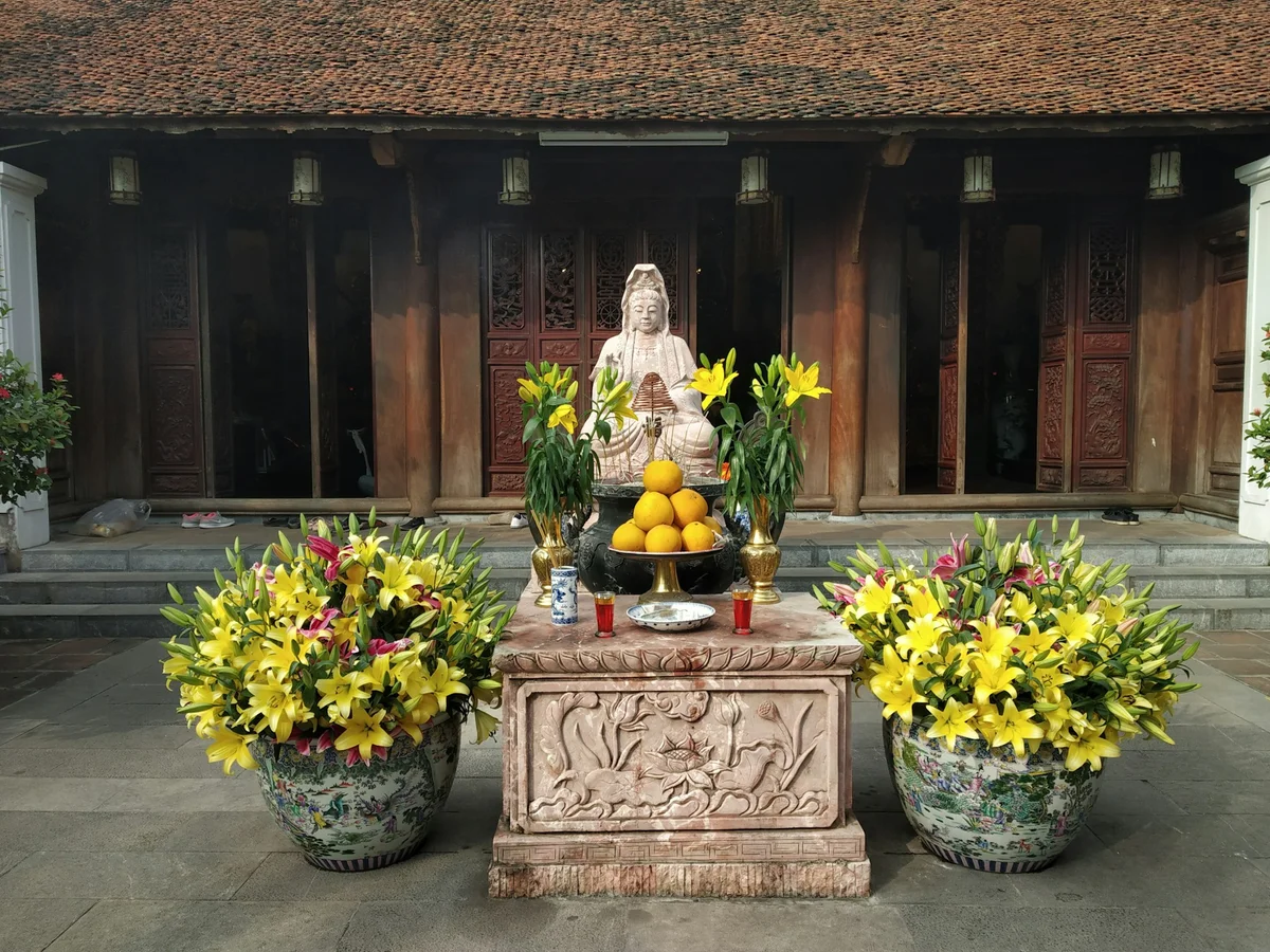 Stone statue surrounded by yellow lilies and fruit offerings on a carved pedestal. Wooden temple with intricate doors in the background.