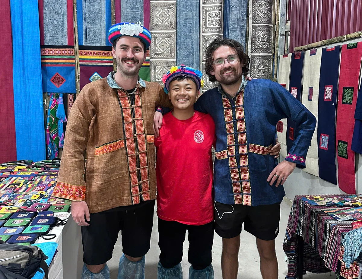 Three people smiling in colorful traditional attire in a vibrant fabric shop. Decorative patterns adorn the walls and tables.