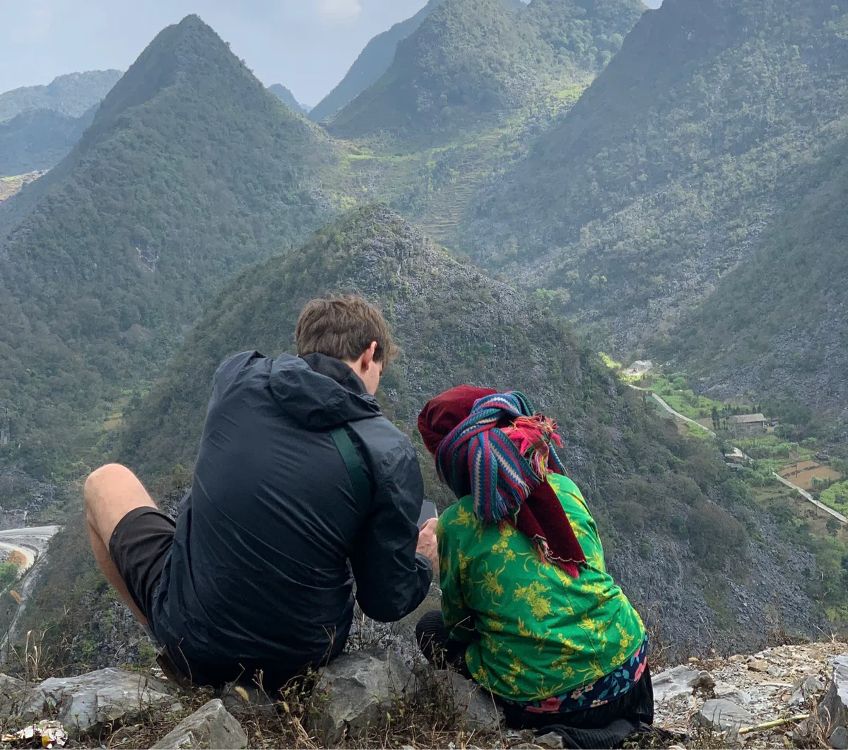 A man and a woman are looking at a phone with the Ha Giang Loop mountains in the background