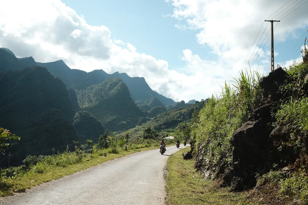 Motorcyclists ride down a rural road flanked by lush greenery and rugged mountains under a bright, partly cloudy sky. Calm atmosphere.