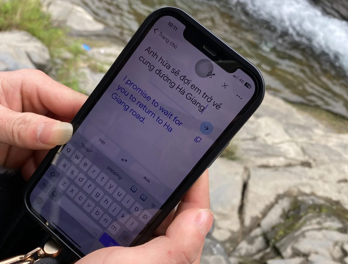 Hands holding a phone displaying a translated message about waiting on Hà Giang road. Background shows rocks and flowing water.
