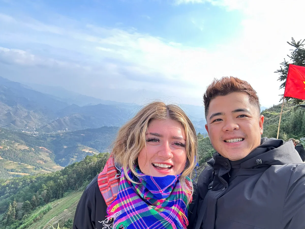 Two smiling people in jackets pose for a selfie on a mountain with a red flag in the background. The sky is clear with scattered clouds.